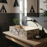 Hand-carved rectangular vessel sink made from Calacatta Amber Viola marble, placed on light wood vanity with matte black fixtures